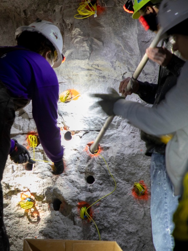 Students inside of cave wearing hardhats with headlamps are putting explosives into drilled holes inside a cave